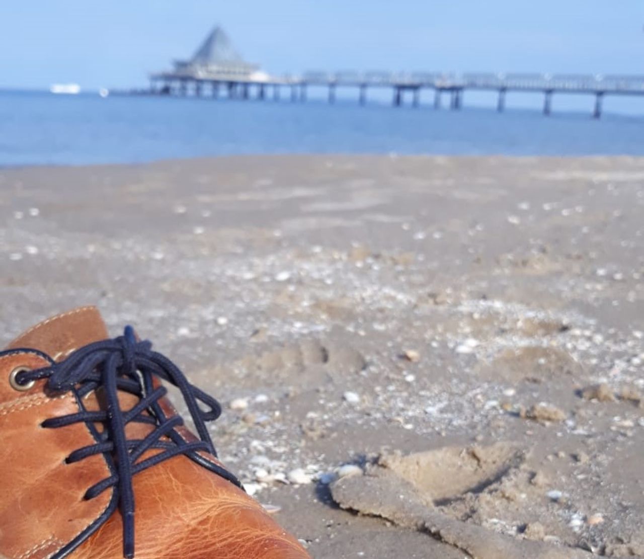 Nahaufnahme von einem Lederschuh am Strand, im Hintergrund laden das Meer und die Seebrücke zum Strandurlaub in Heringsdorf ein